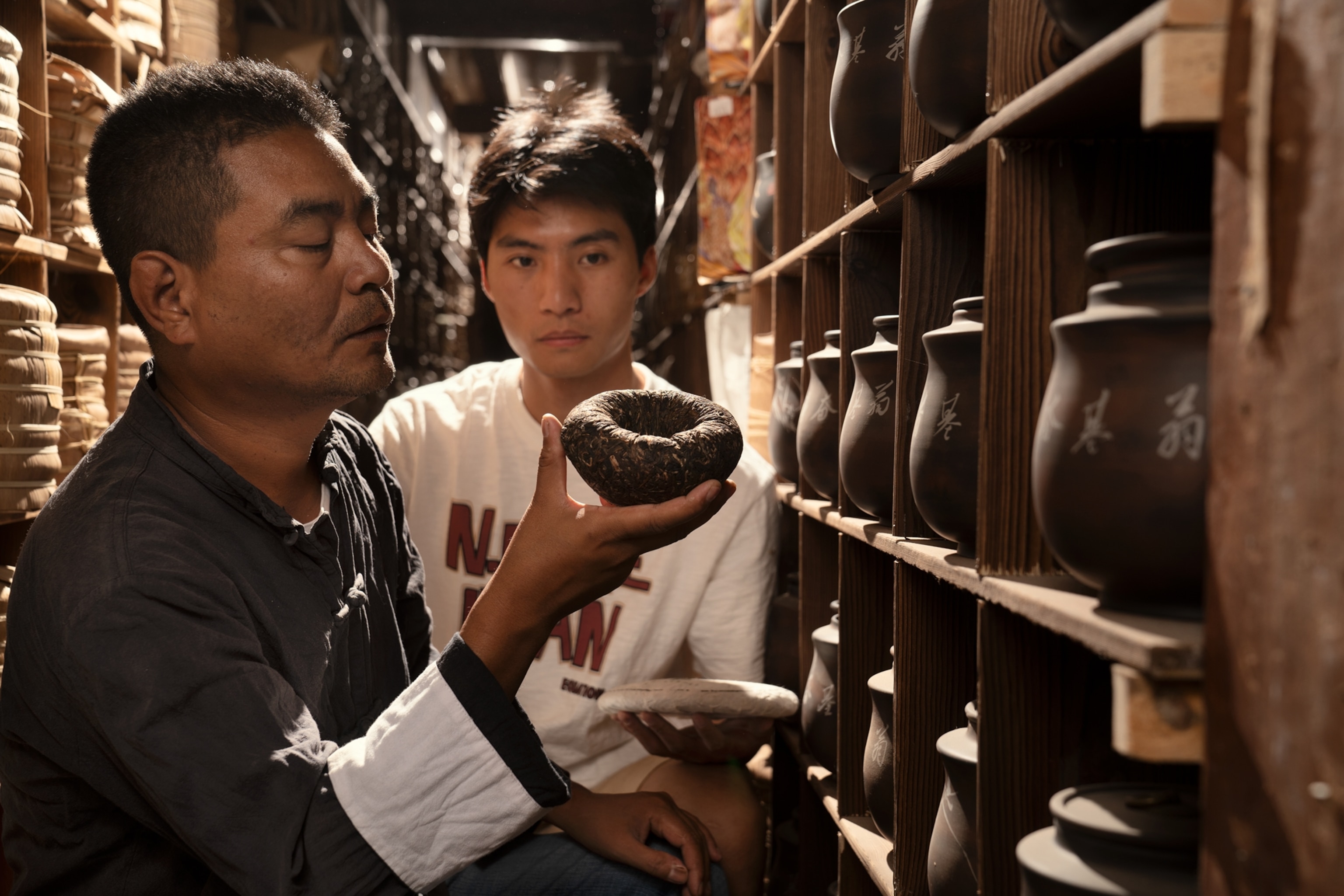 A tea maker and apprentice inspecting aging cakes in a timber storeroom
